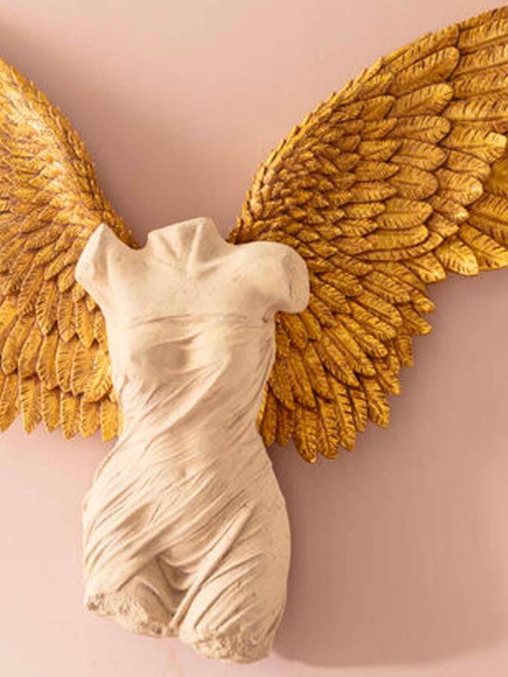 A large sculpture featuring majestic, highly detailed gold wings that shimmer with a metallic finish. The wings extend gracefully upwards and outwards, resembling an angelic or mythical creature. The central figure is a white female form with smooth, marble-like features, exuding elegance and serenity. Her posture is poised, as though in flight or ascending, with her arms reaching gracefully toward the heavens or softly at her sides. The base of the statue is minimalistic, perhaps a sleek metallic or stone 